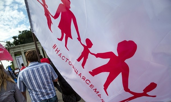 A person pictured from behind carrying a large white flag with an outline of children and adults holding hands and Cyrillic text underneath.