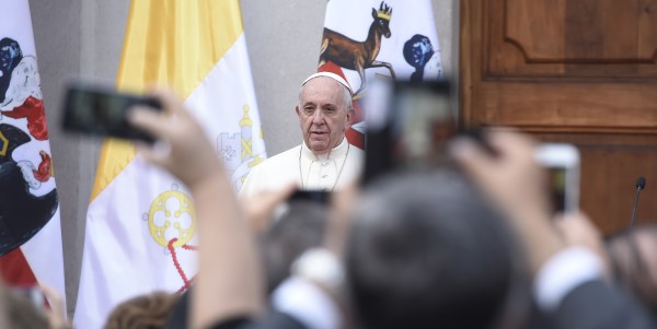 Pope Francis addressing a crowd in focus in the background with raised arms taking pictures of him in the foreground