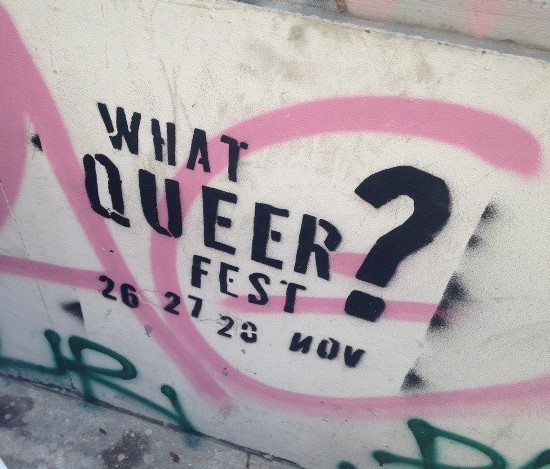the text 'what queer fest?' sprayed in black on a concrete wall in black along with pink and green graffiti